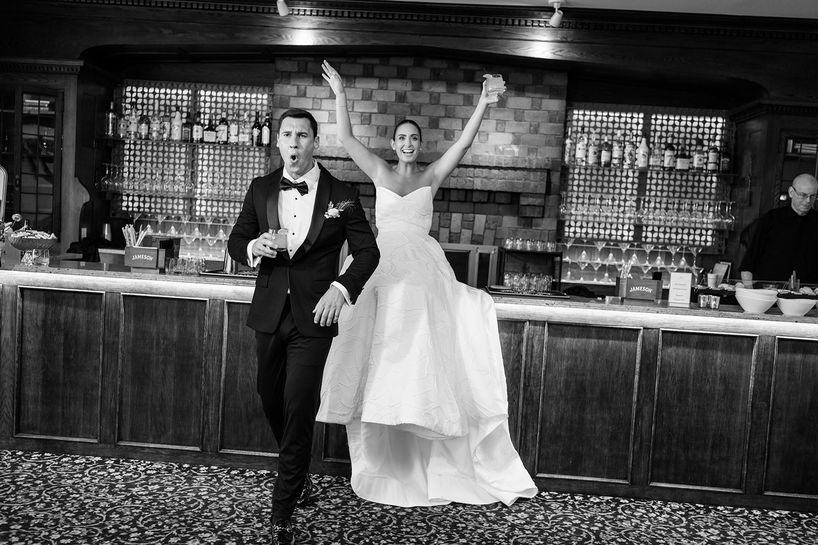 Black and white film photo of the bride and groom cheering playfully with drinks at the bar.